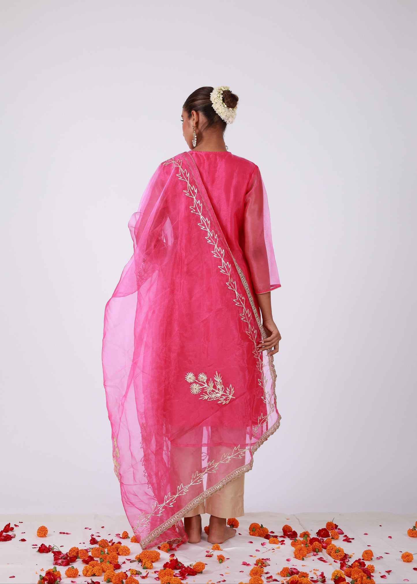 Model wearing a rani pink organza kurta set with gold embroidery and matching organza dupatta, paired with light gold silk pants – standing on a floral setup.
