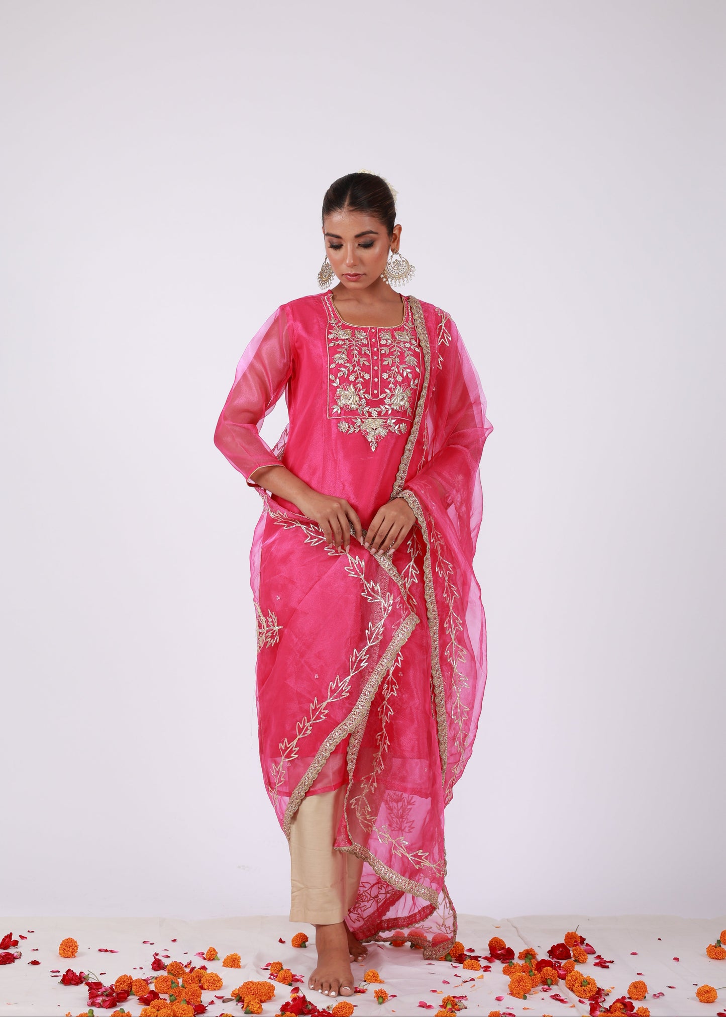 Model wearing a rani pink organza kurta set with gold embroidery and matching organza dupatta, paired with light gold silk pants – standing on a floral setup.