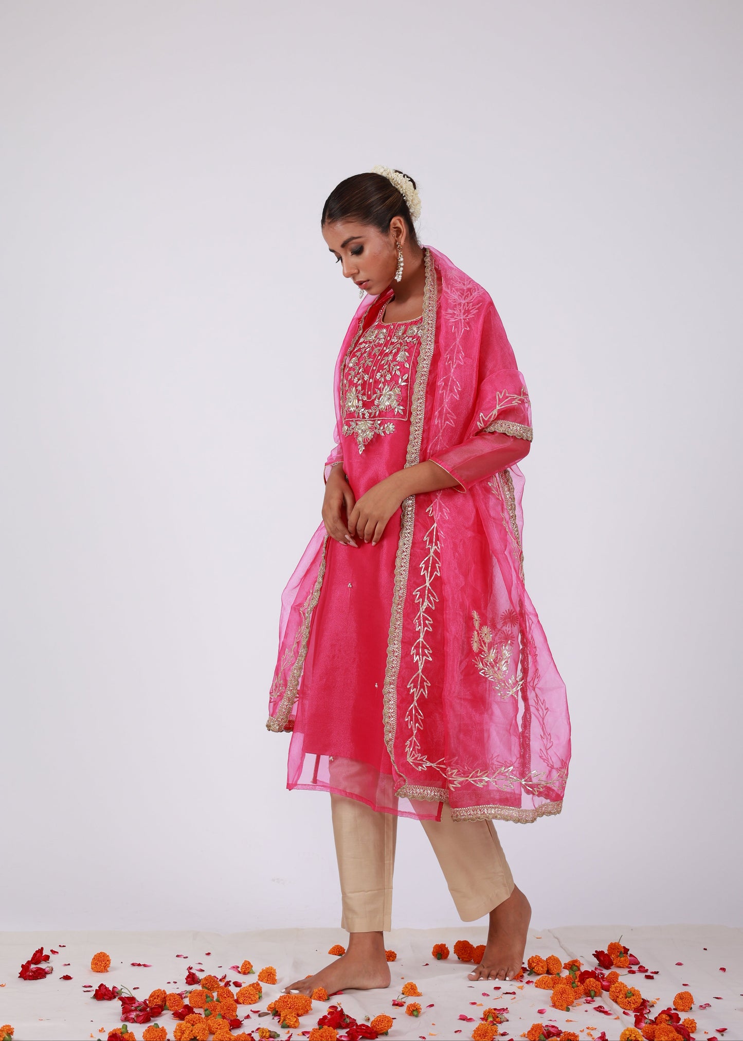 Model wearing a rani pink organza kurta set with gold embroidery and matching organza dupatta, paired with light gold silk pants – standing on a floral setup.