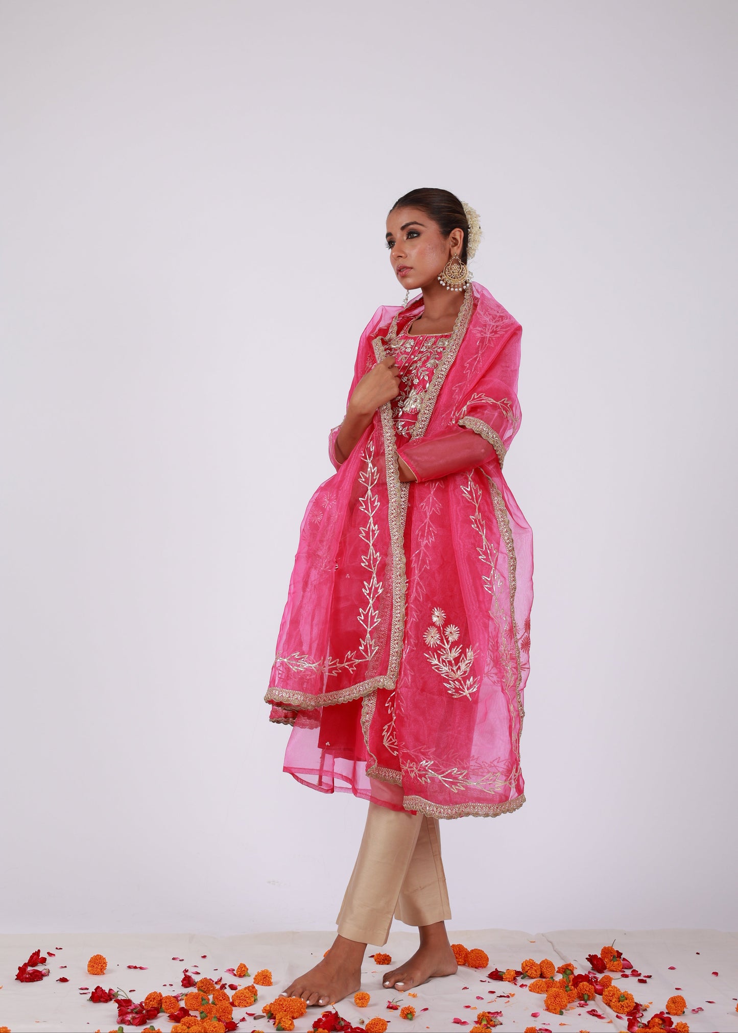 Model wearing a rani pink organza kurta set with gold embroidery and matching organza dupatta, paired with light gold silk pants – standing on a floral setup.