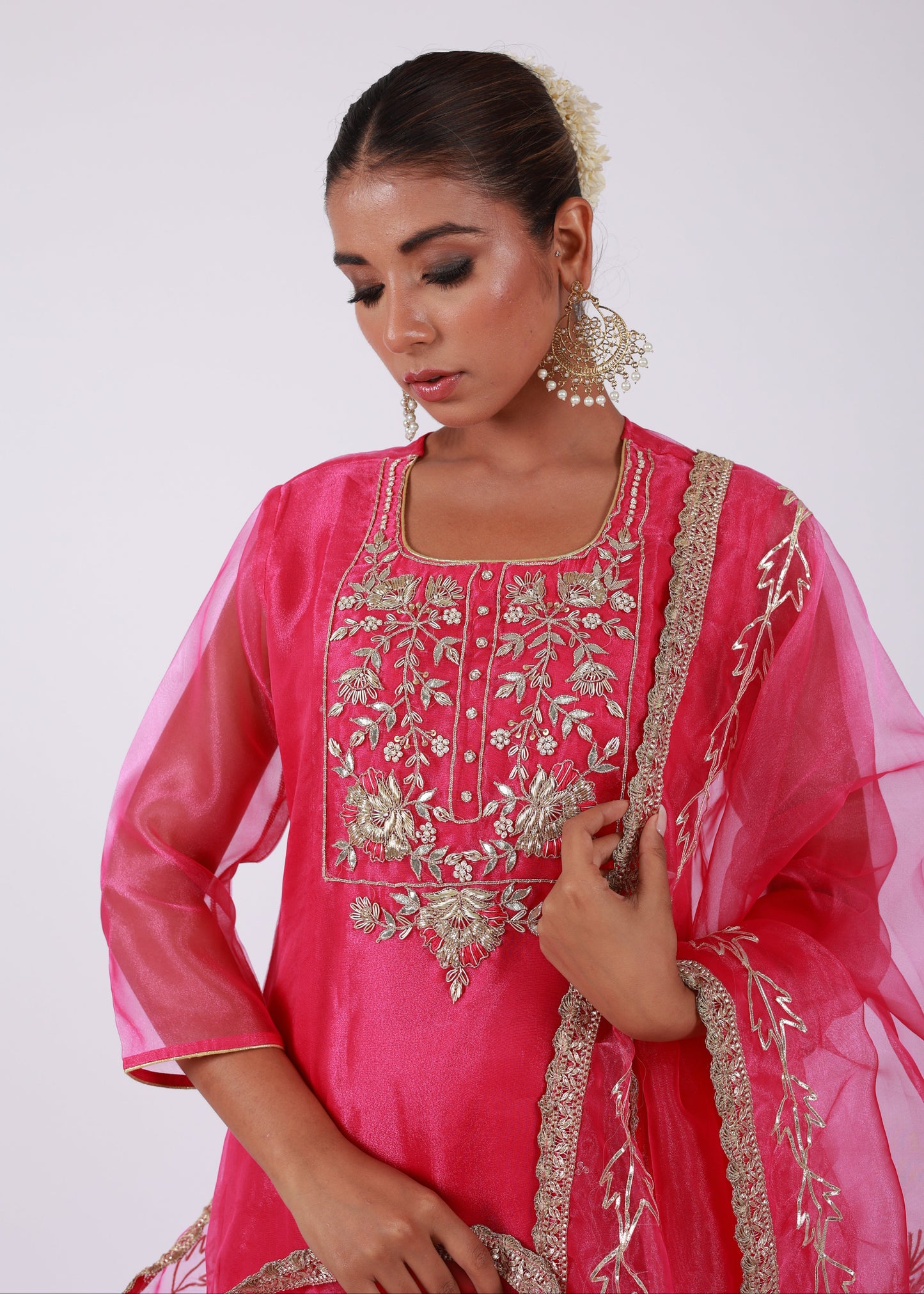 Model wearing a rani pink organza kurta set with gold embroidery and matching organza dupatta, paired with light gold silk pants – standing on a floral setup.