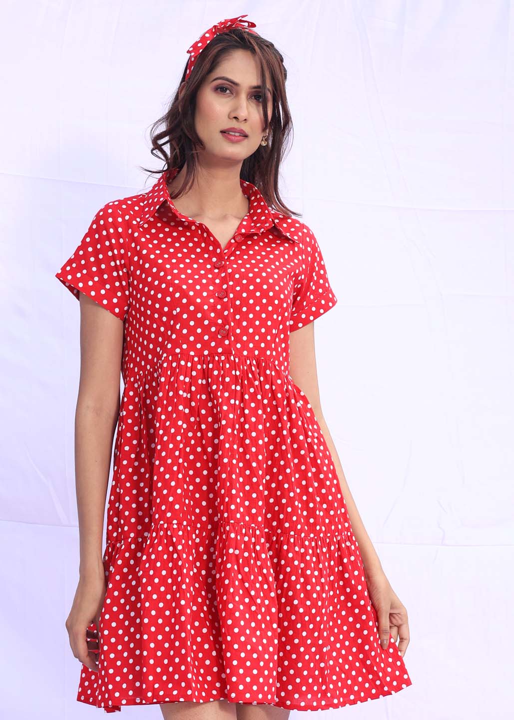 Woman wearing a red cotton flared dress with white polka dots and a collared neckline, styled with a matching red hair bow, against a soft lavender backdrop.