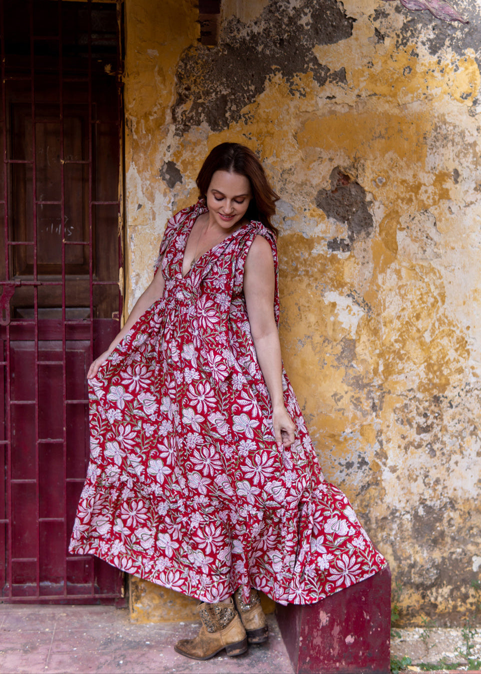 Woman wearing a red floral handblock printed cotton maxi dress with tie-knot shoulders and deep V-neck, standing against a rustic yellow wall.