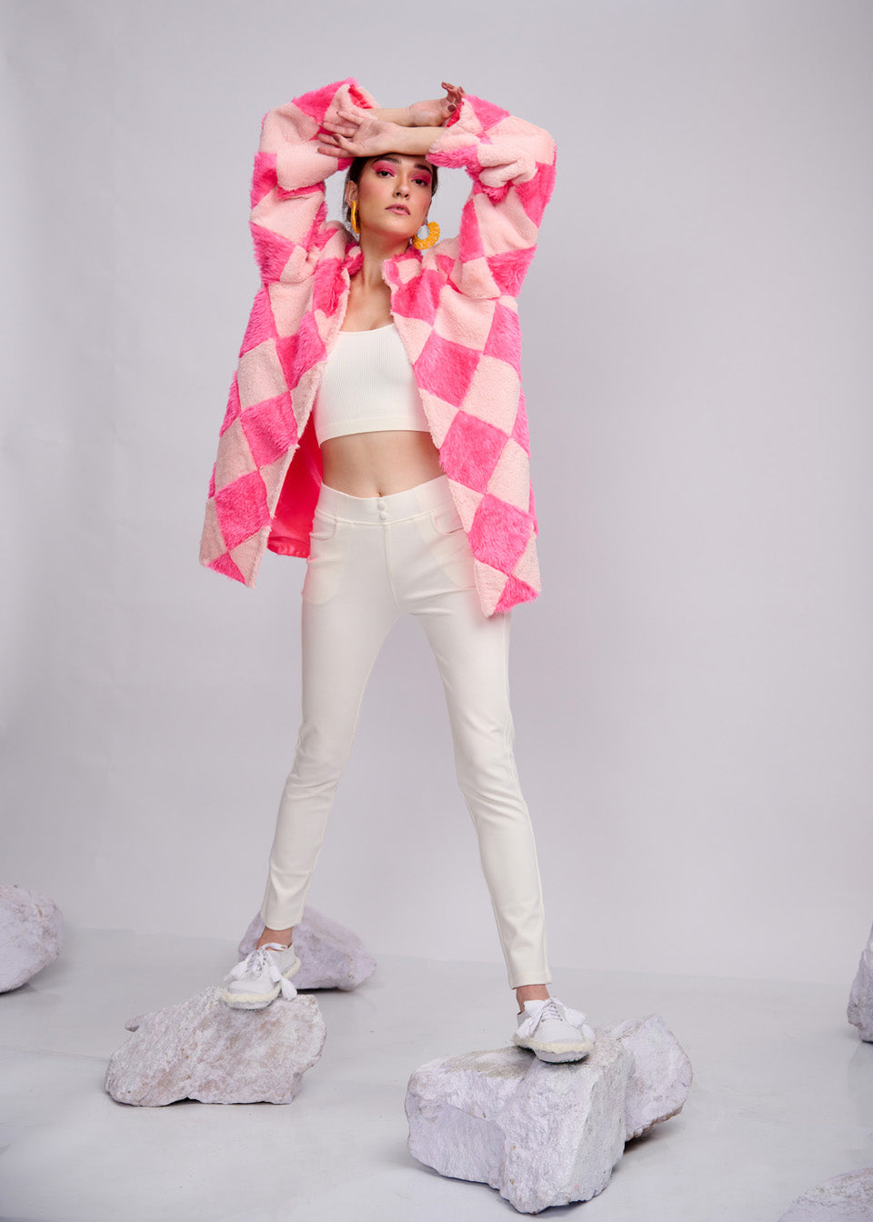 Model wearing a vibrant pink and blush faux fur checkerboard jacket from The Silver Lining collection, styled with white pants and crop top, standing confidently on marble-like stones.