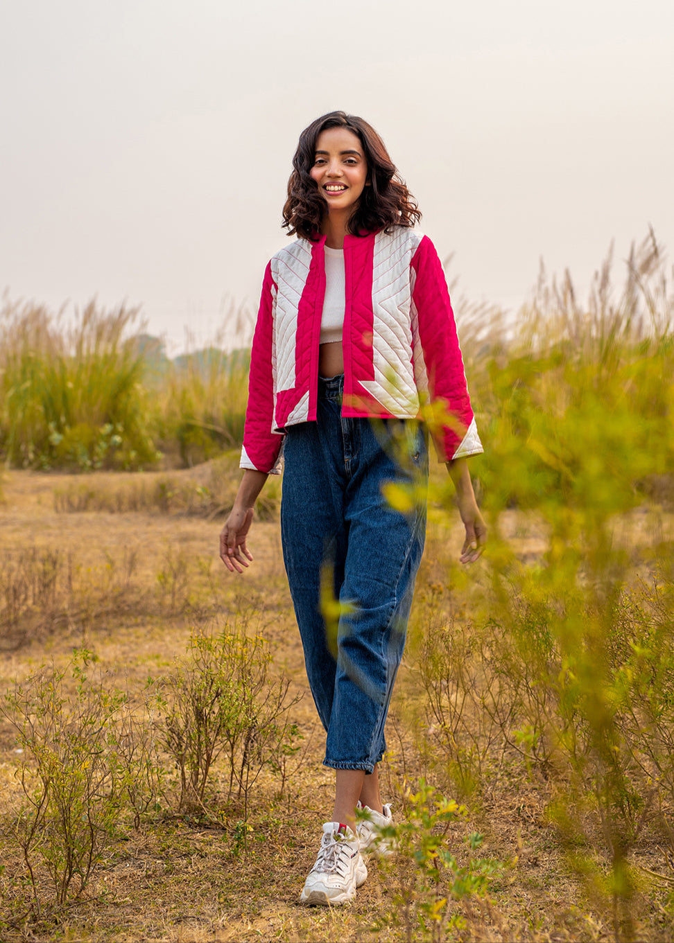 A young woman wearing a pink and white patchwork quilted jacket styled with high-waist blue jeans and a white crop top, smiling outdoors in a dry grass field.