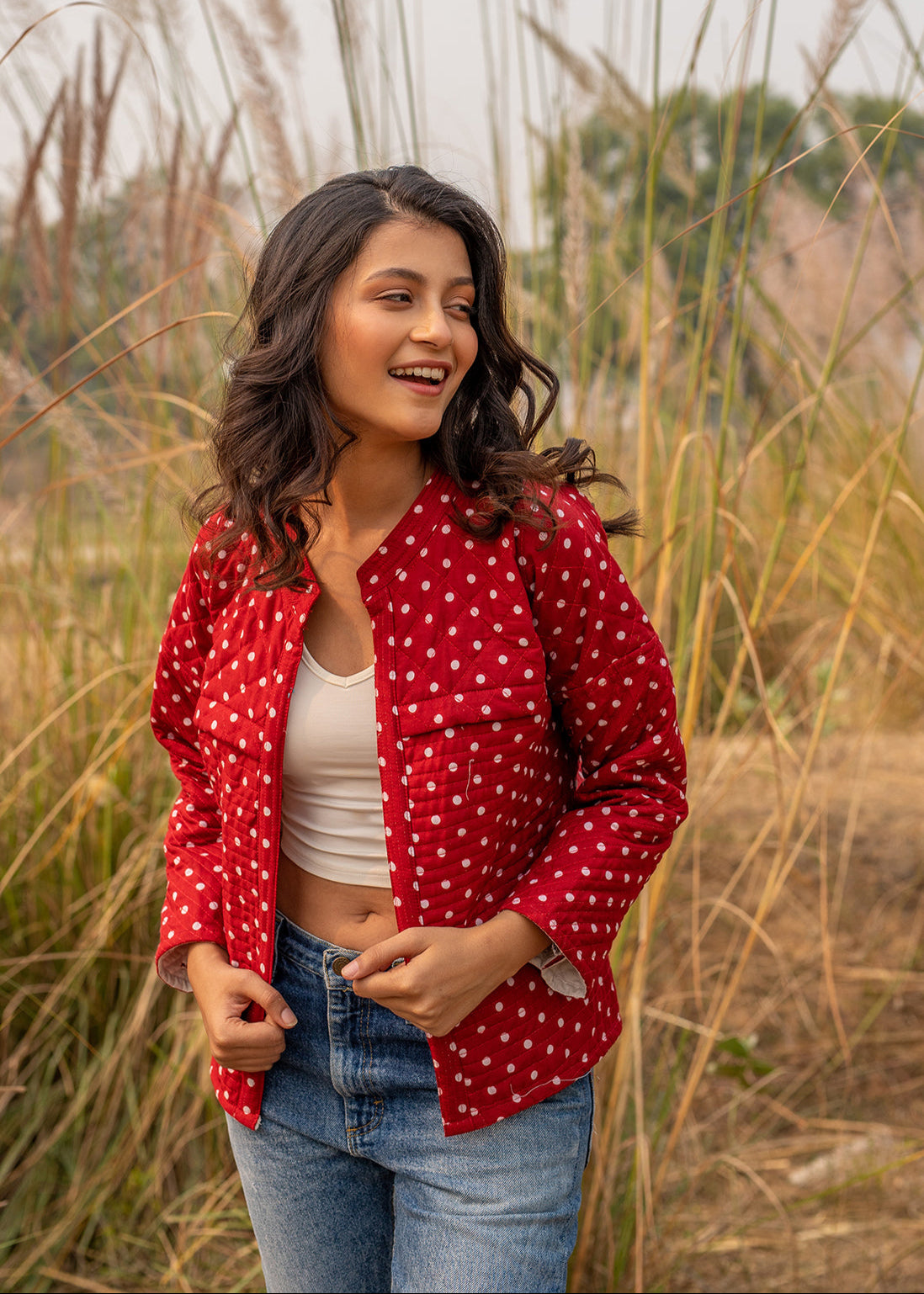 Woman in a maroon polka dot quilted cotton jacket styled over a white crop top and denim, smiling in an outdoor setting with tall grass in the background.