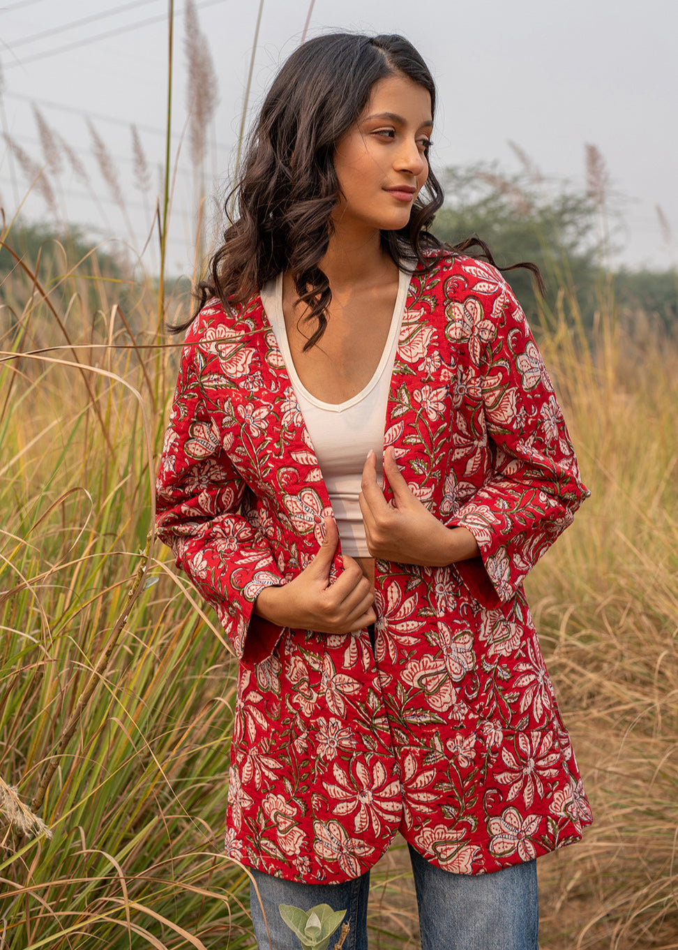 Woman wearing a red hand block printed floral jacket over a white crop top and denim, standing in a grassy field with a serene expression.