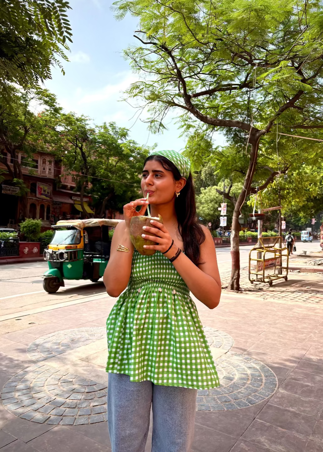 Woman in a green checkered LilacLoops - smocked tops That Romeo drinking from a cup on a street with trees and a rickshaw in the background.