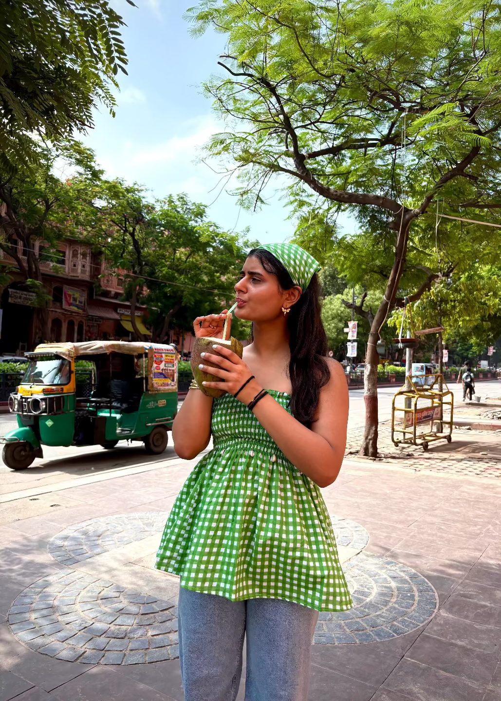 Woman in a green checkered LilacLoops - smocked tops That Romeo drinking from a cup on a street with trees and a rickshaw in the background.