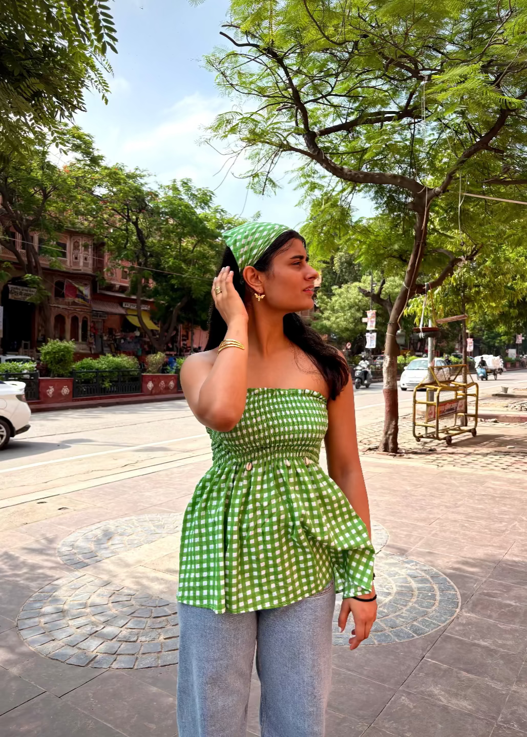 Woman wearing a green checkered LilacLoops - smocked tops That Romeo top and light blue jeans standing on a street with trees and buildings in the background.