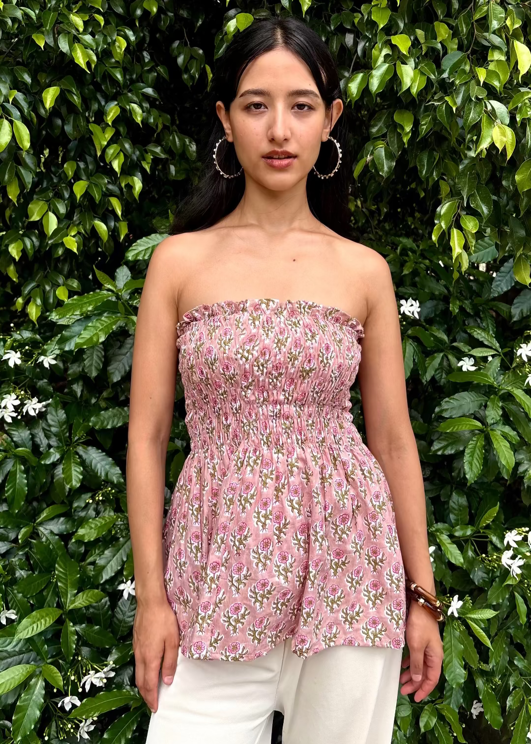 Woman wearing a pink strapless PuffBindi- smocked tops That Romeo dress standing in front of green foliage