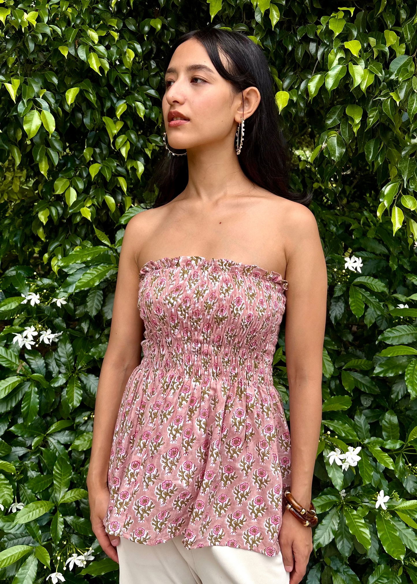 Woman wearing a pink strapless PuffBindi- smocked tops That Romeo dress standing in front of green foliage