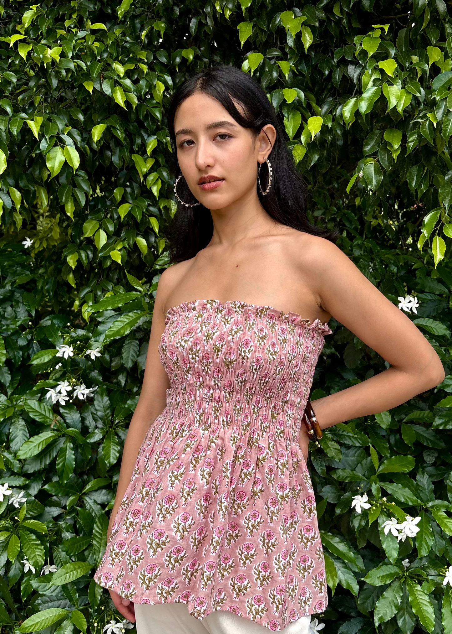 Woman wearing a pink strapless PuffBindi- smocked tops That Romeo dress standing in front of green foliage