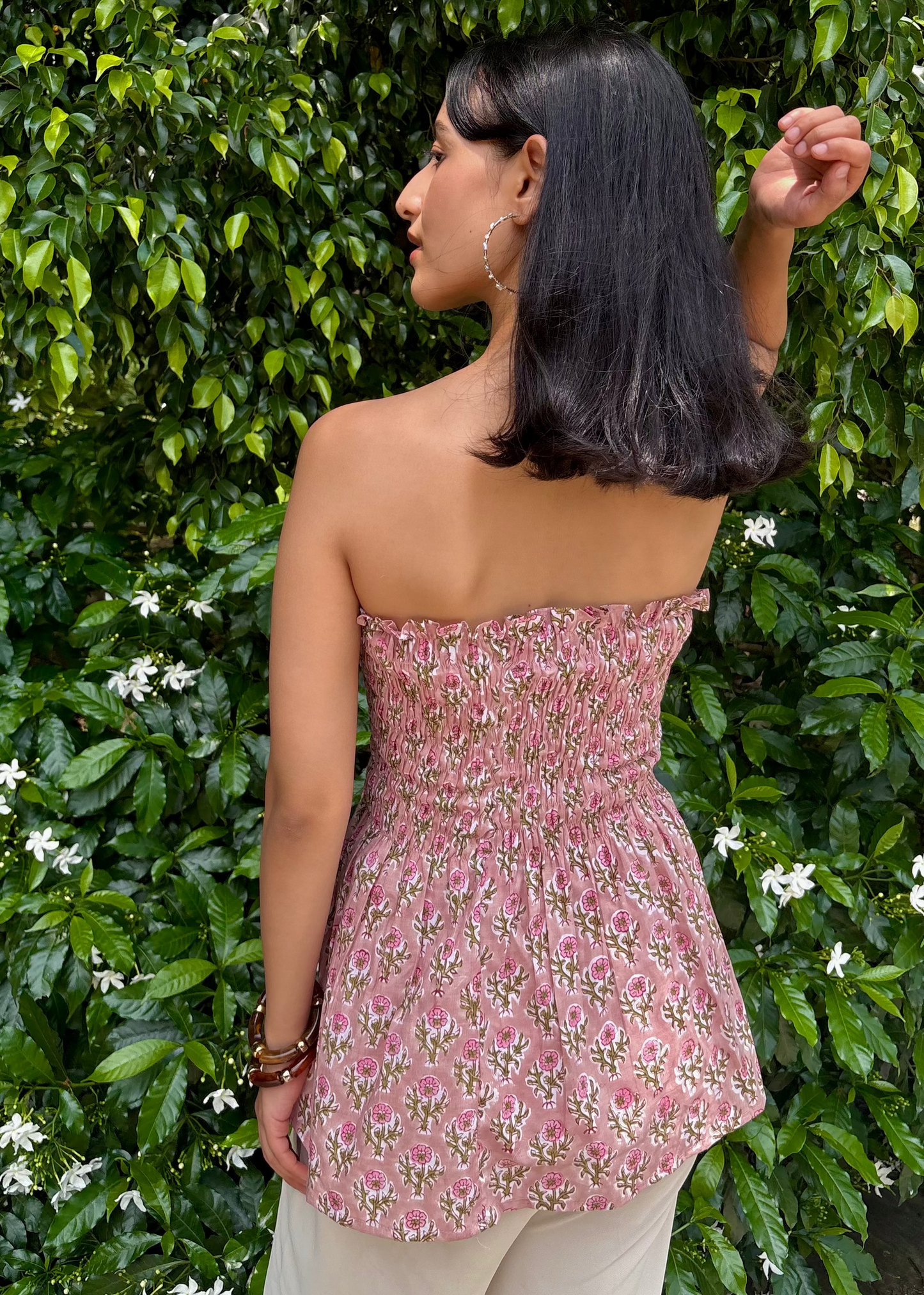 Woman wearing a pink strapless PuffBindi- smocked tops That Romeo dress standing in front of green foliage