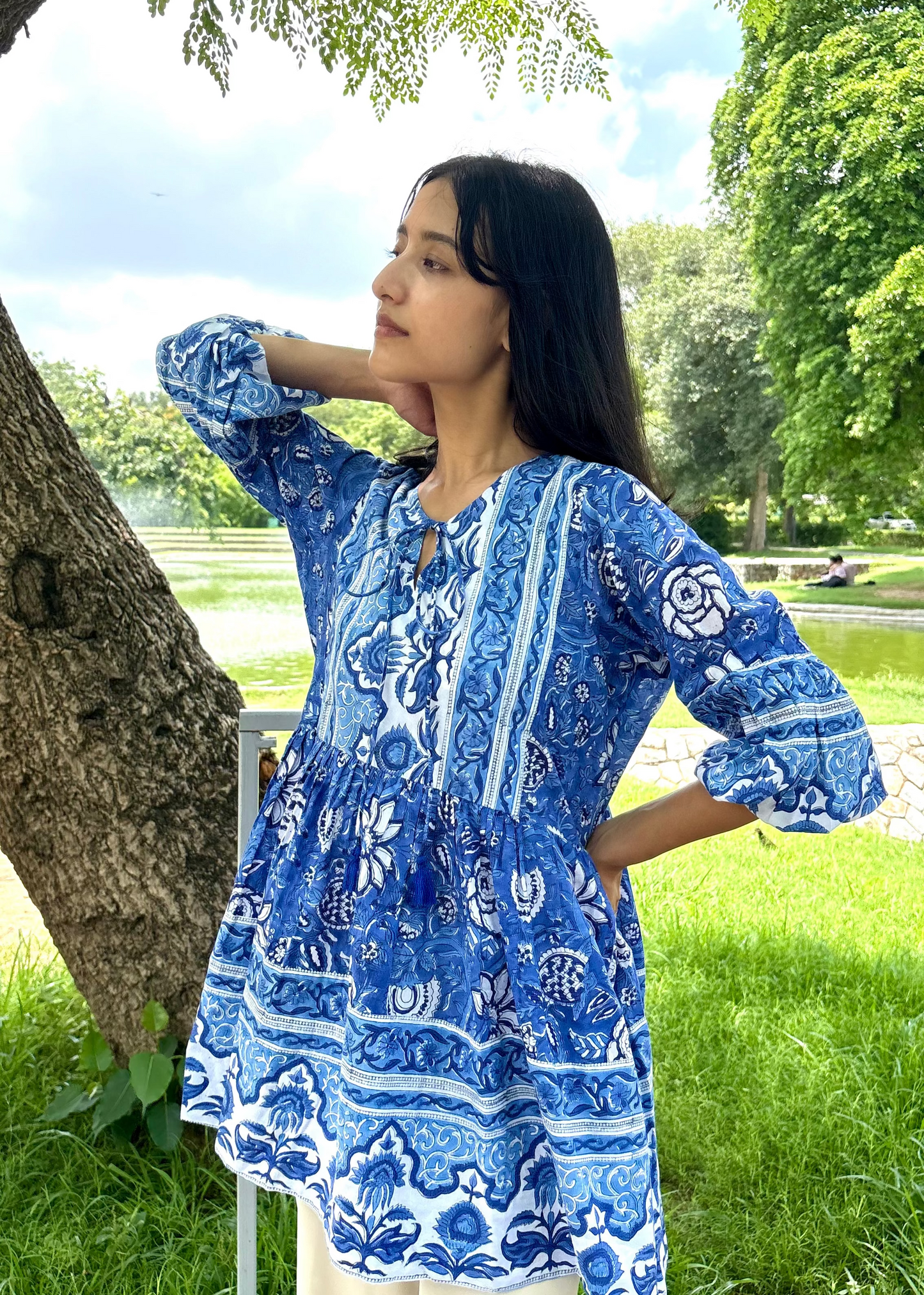 Woman wearing a blue and white patterned Floral Whispers Tunic top That Romeo dress standing outdoors near a tree and body of water.