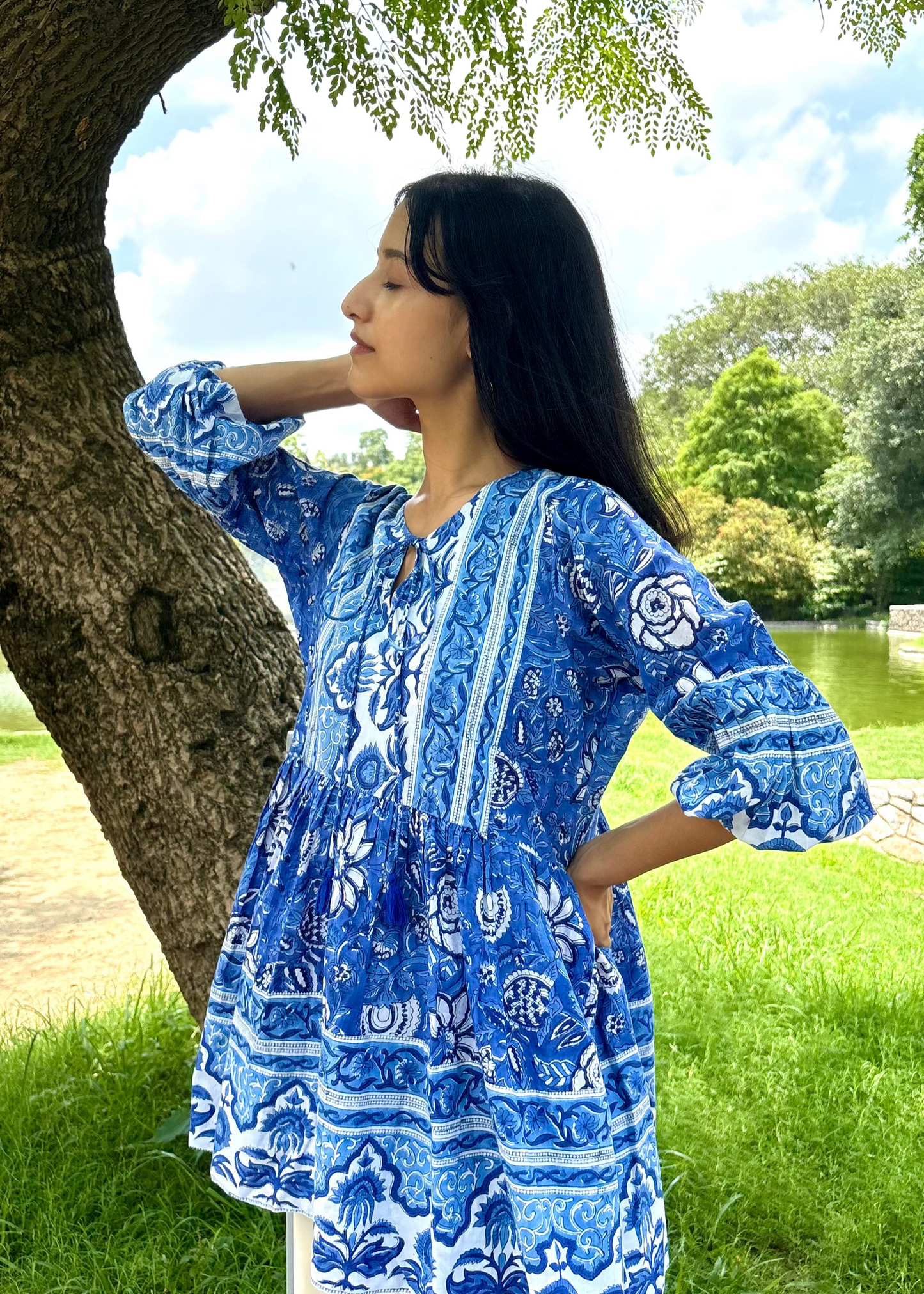 Woman wearing a blue and white patterned Floral Whispers Tunic top That Romeo dress standing outdoors near a tree and body of water.