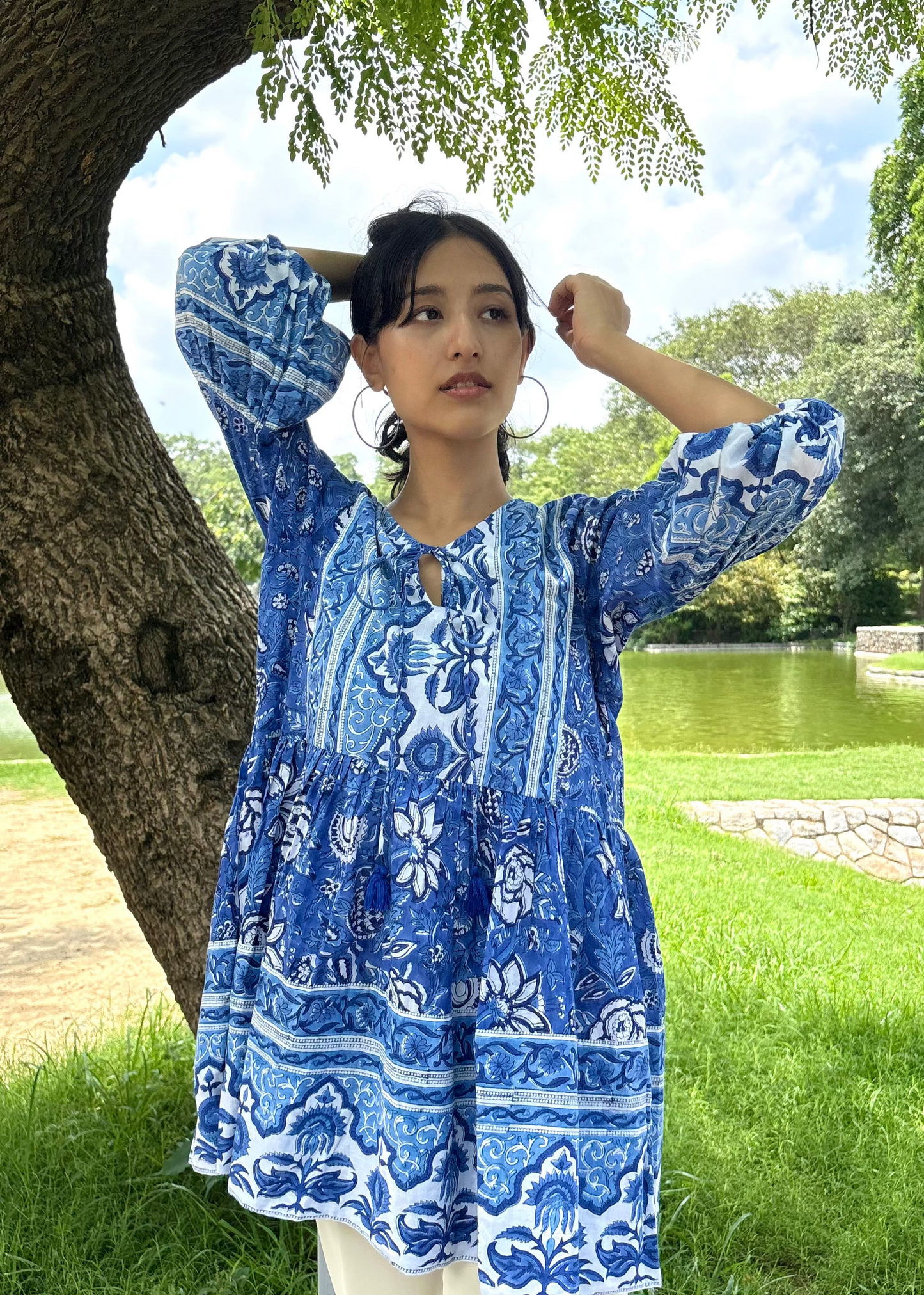 Woman wearing a blue and white patterned Floral Whispers Tunic top That Romeo dress standing outdoors near a tree and body of water.