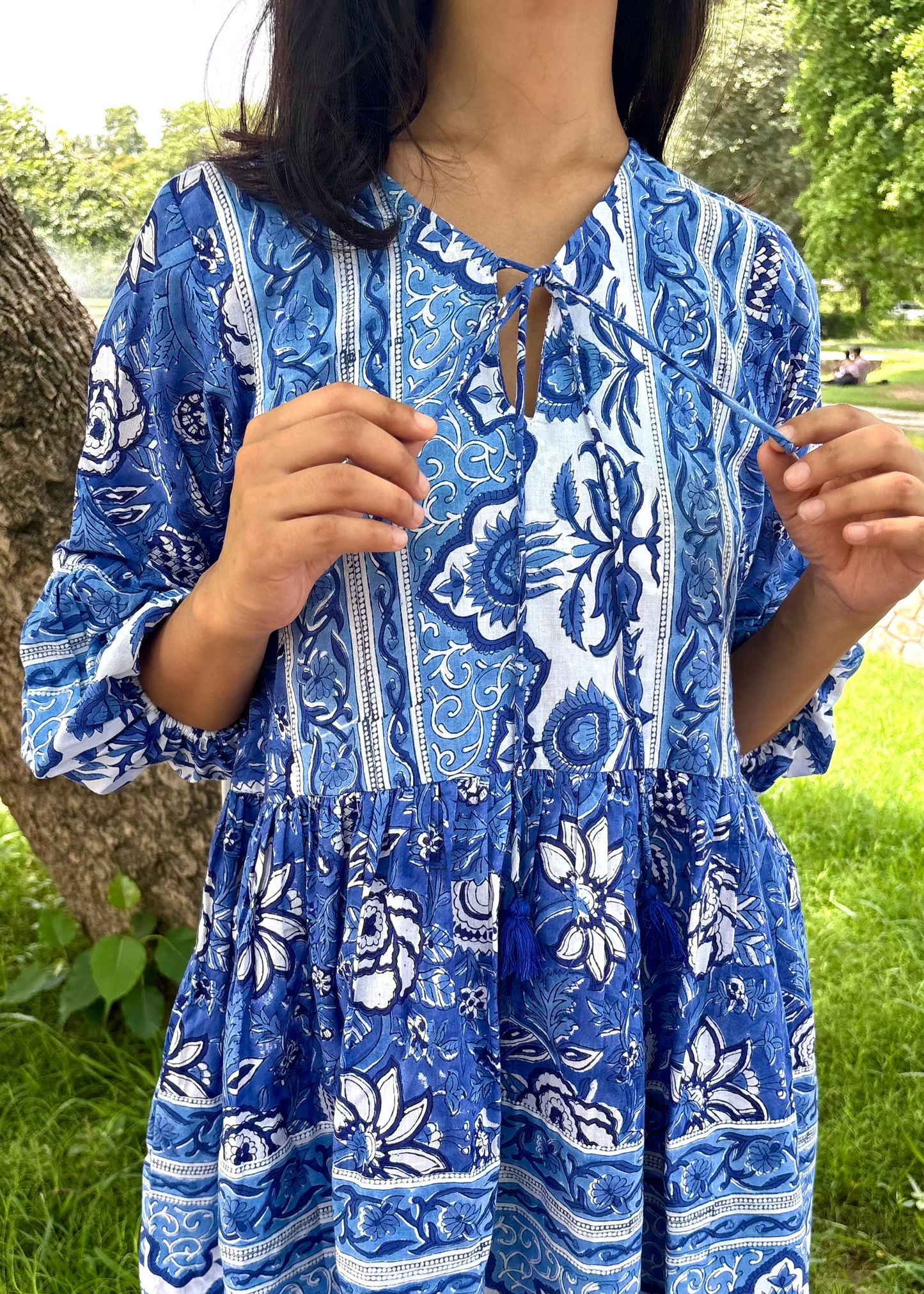 Woman wearing a blue and white patterned Floral Whispers Tunic top That Romeo dress standing outdoors near a tree and body of water.