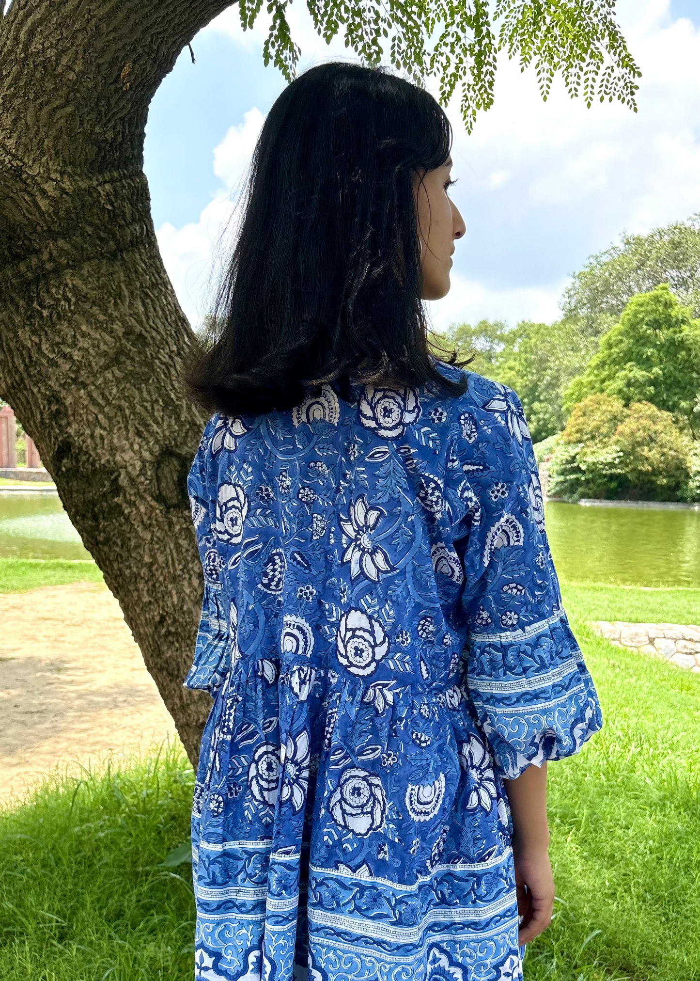 Woman wearing a blue and white patterned Floral Whispers Tunic top That Romeo dress standing outdoors near a tree and body of water.