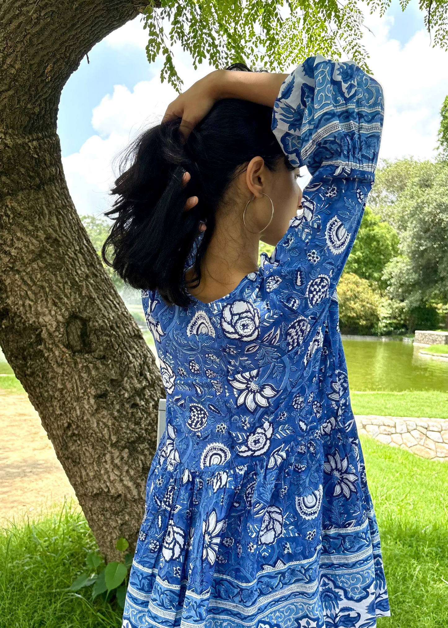 Woman wearing a blue and white patterned Floral Whispers Tunic top That Romeo dress standing outdoors near a tree and body of water.