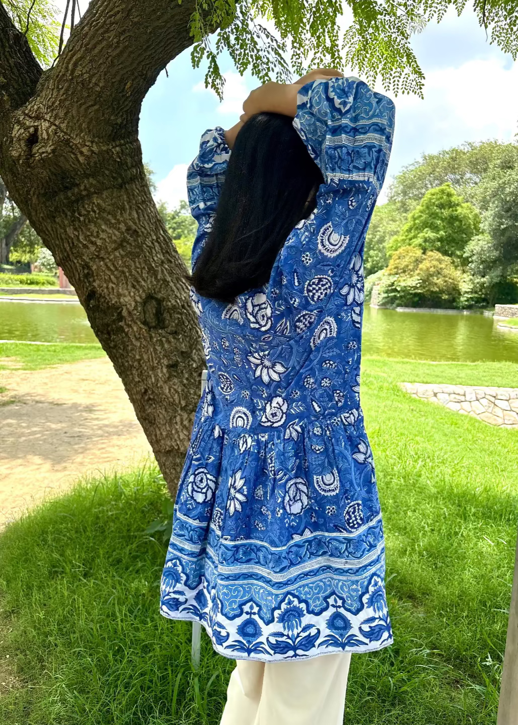 Woman wearing a blue and white patterned Floral Whispers Tunic top That Romeo dress standing outdoors near a tree and body of water.