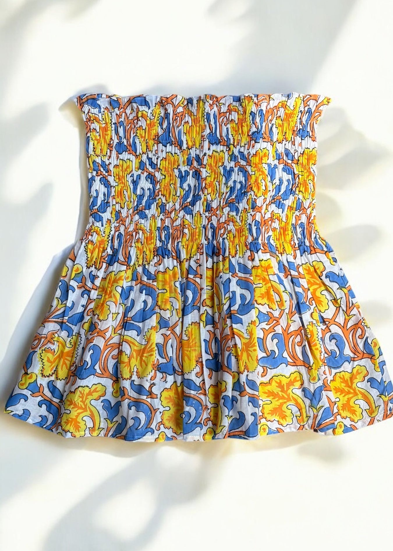 A strapless cotton smocked top with a bold yellow, blue, and orange baroque floral print, flared below the bust and laid flat under soft shadows by That Romeo