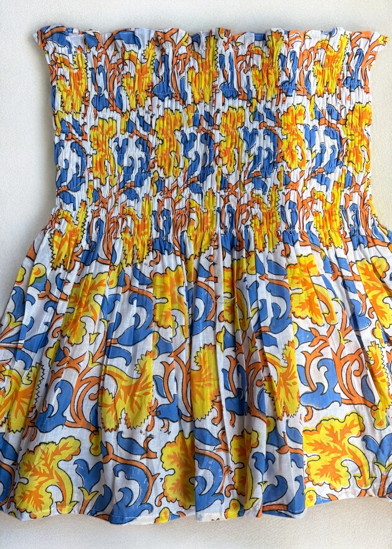 A strapless cotton smocked top with a bold yellow, blue, and orange baroque floral print, flared below the bust and laid flat under soft shadows by That Romeo