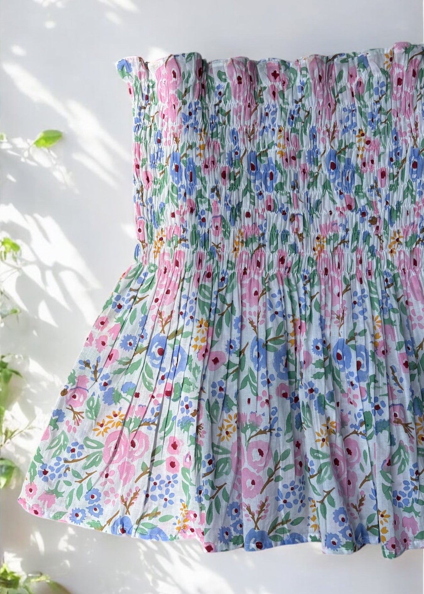 A strapless floral smocked top in pastel shades of pink, blue, and green, flared below the bust and photographed against a softly lit wall with leafy shadows by That Romeo