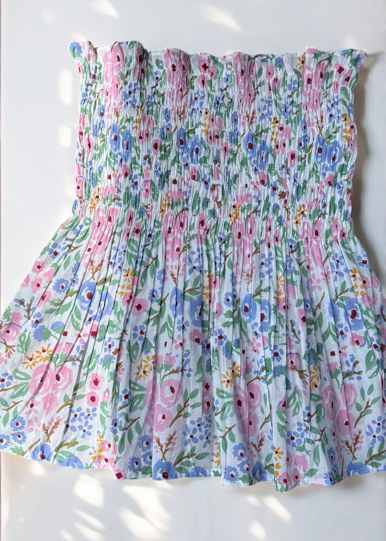 A strapless floral smocked top in pastel shades of pink, blue, and green, flared below the bust and photographed against a softly lit wall with leafy shadows by That Romeo