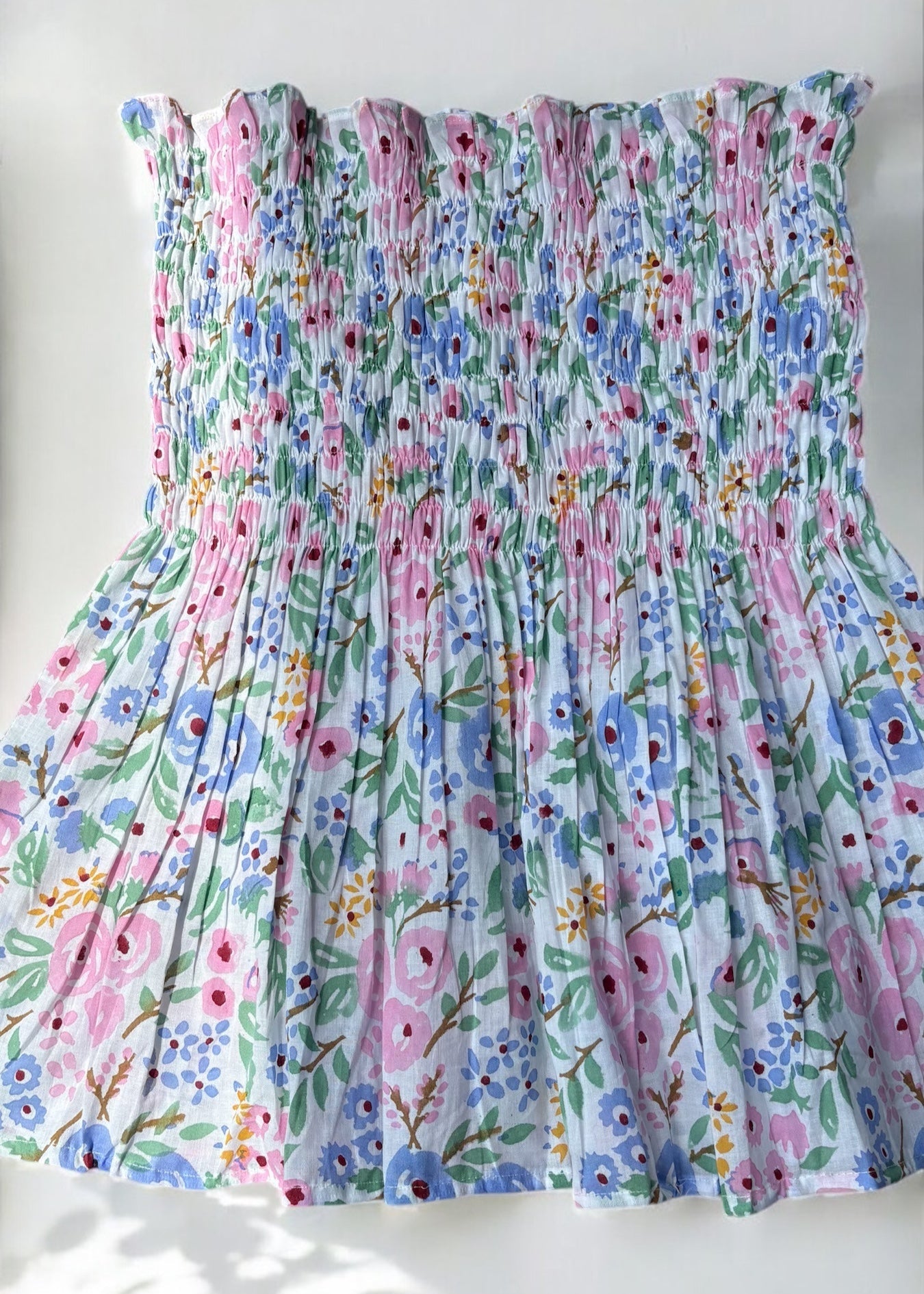 A strapless floral smocked top in pastel shades of pink, blue, and green, flared below the bust and photographed against a softly lit wall with leafy shadows by That Romeo