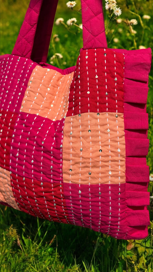 A quilted tote bag with vertical Kantha stitch embroidery in rose, berry, and coral shades, featuring ruffled edges.