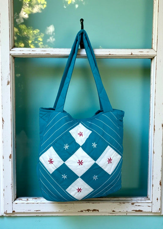 Teal quilted cotton tote bag with white patchwork panel and embroidered floral motifs hanging on a window frame.