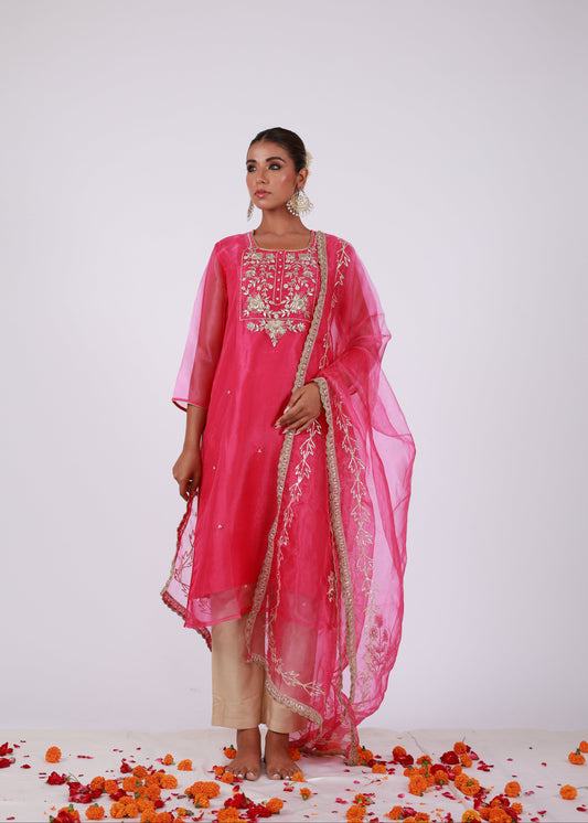 Model wearing a rani pink organza kurta set with gold embroidery and matching organza dupatta, paired with light gold silk pants – standing on a floral setup.