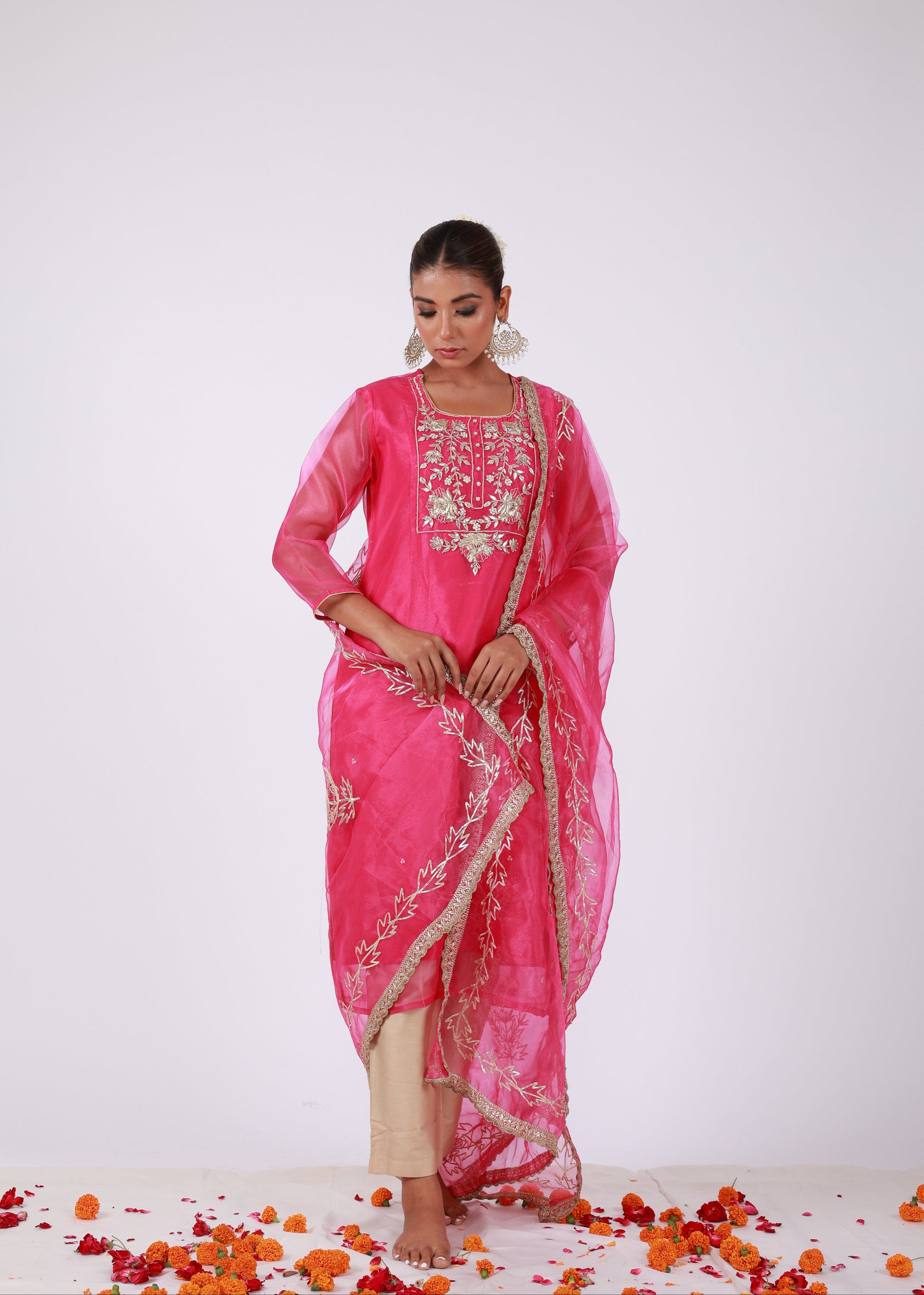 Model wearing a rani pink organza kurta set with gold embroidery and matching organza dupatta, paired with light gold silk pants – standing on a floral setup.
