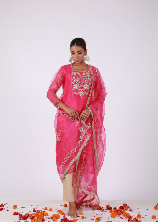 Model wearing a rani pink organza kurta set with gold embroidery and matching organza dupatta, paired with light gold silk pants – standing on a floral setup.