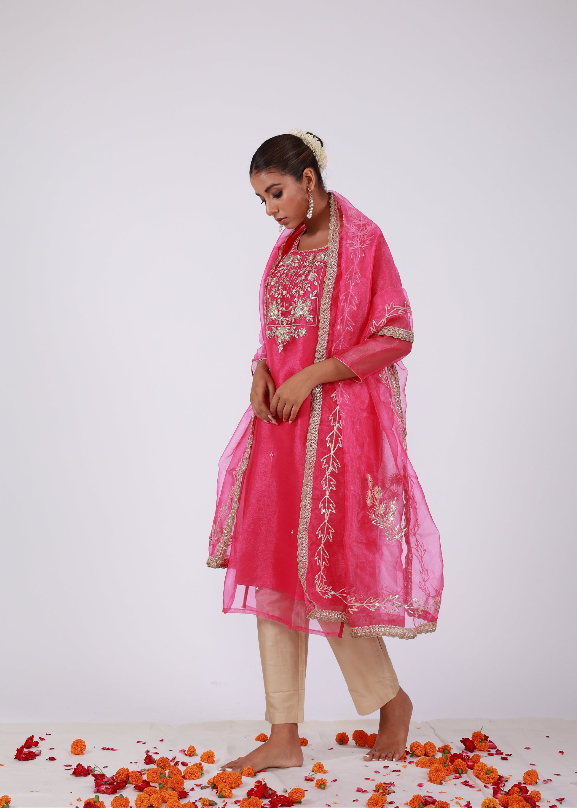 Model wearing a rani pink organza kurta set with gold embroidery and matching organza dupatta, paired with light gold silk pants – standing on a floral setup.