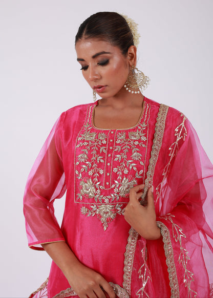 Model wearing a rani pink organza kurta set with gold embroidery and matching organza dupatta, paired with light gold silk pants – standing on a floral setup.