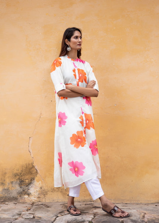 Model wearing a white Chanderi silk kurta with oversized pink and orange floral print, paired with white cotton pants — against a warm ochre wall.

