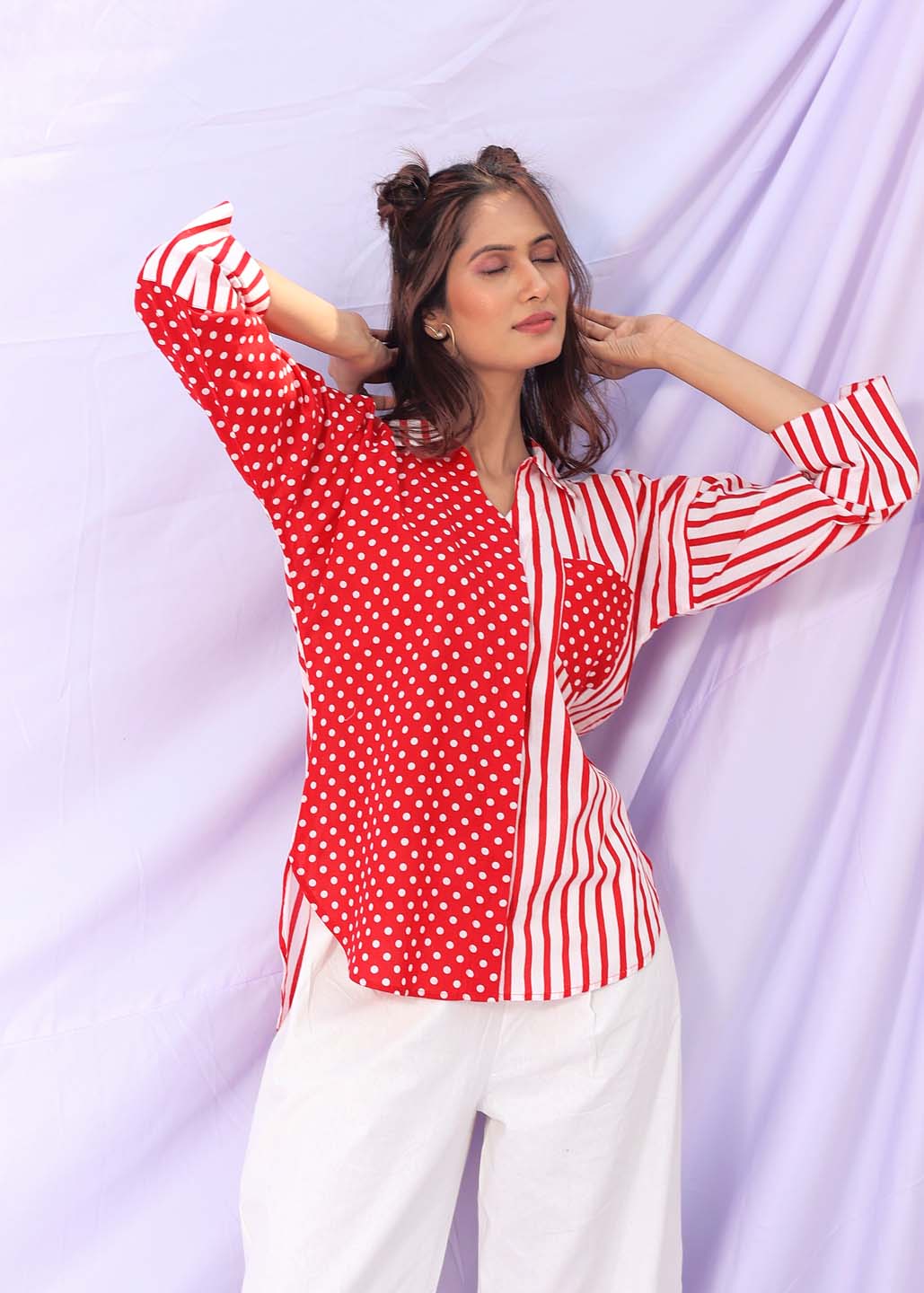 Woman wearing a red cotton shirt with one half polka-dotted and the other half striped, paired with white pants, posing against a pastel lavender backdrop.

