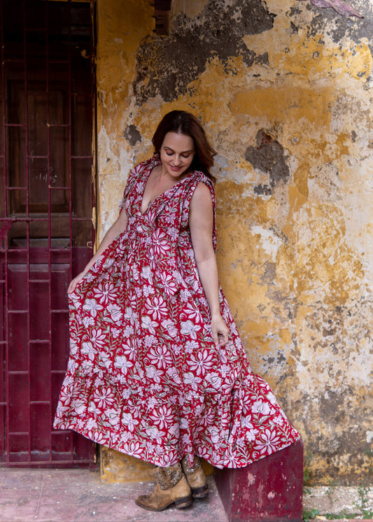Woman wearing a red floral handblock printed cotton maxi dress with tie-knot shoulders and deep V-neck, standing against a rustic yellow wall.