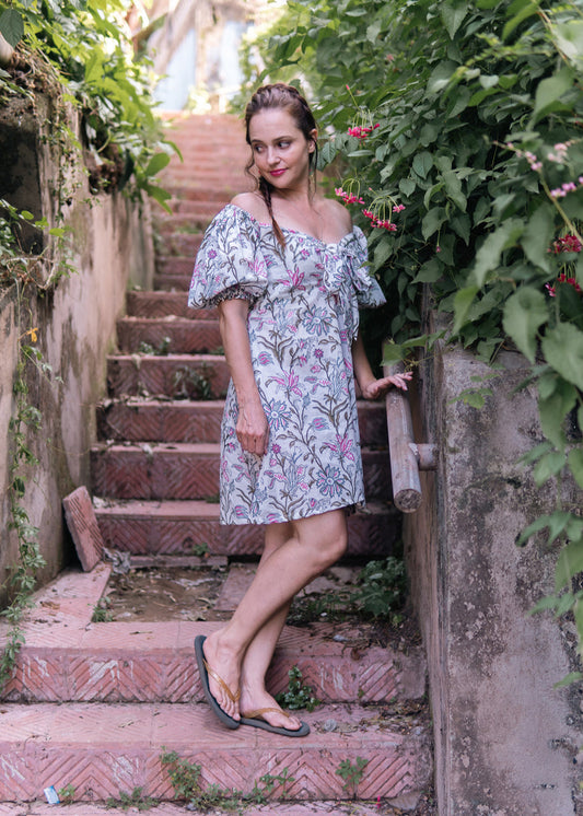 Woman wearing a short off-shoulder hand block printed floral dress with balloon sleeves, standing on rustic stairs surrounded by lush greenery, looking serene and carefree.

