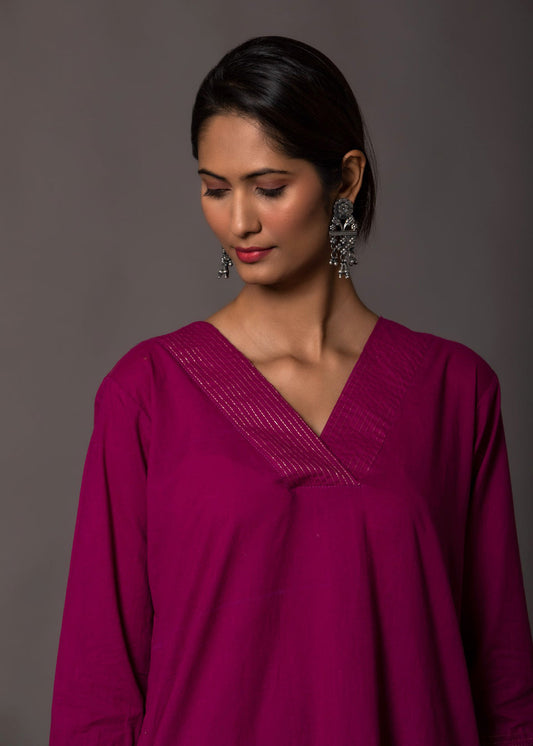 Woman wearing a magenta and pink A-line kurta with gold gota work and matching pants, styled with silver earrings and juttis, against a grey backdrop.

