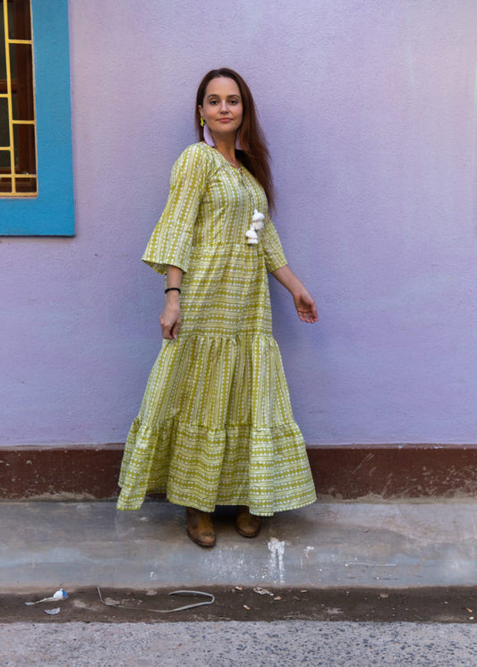 Woman wearing a lime green hand block printed cotton maxi dress with bell sleeves, tassel detailing, and a tiered hem, standing against a pastel purple wall.