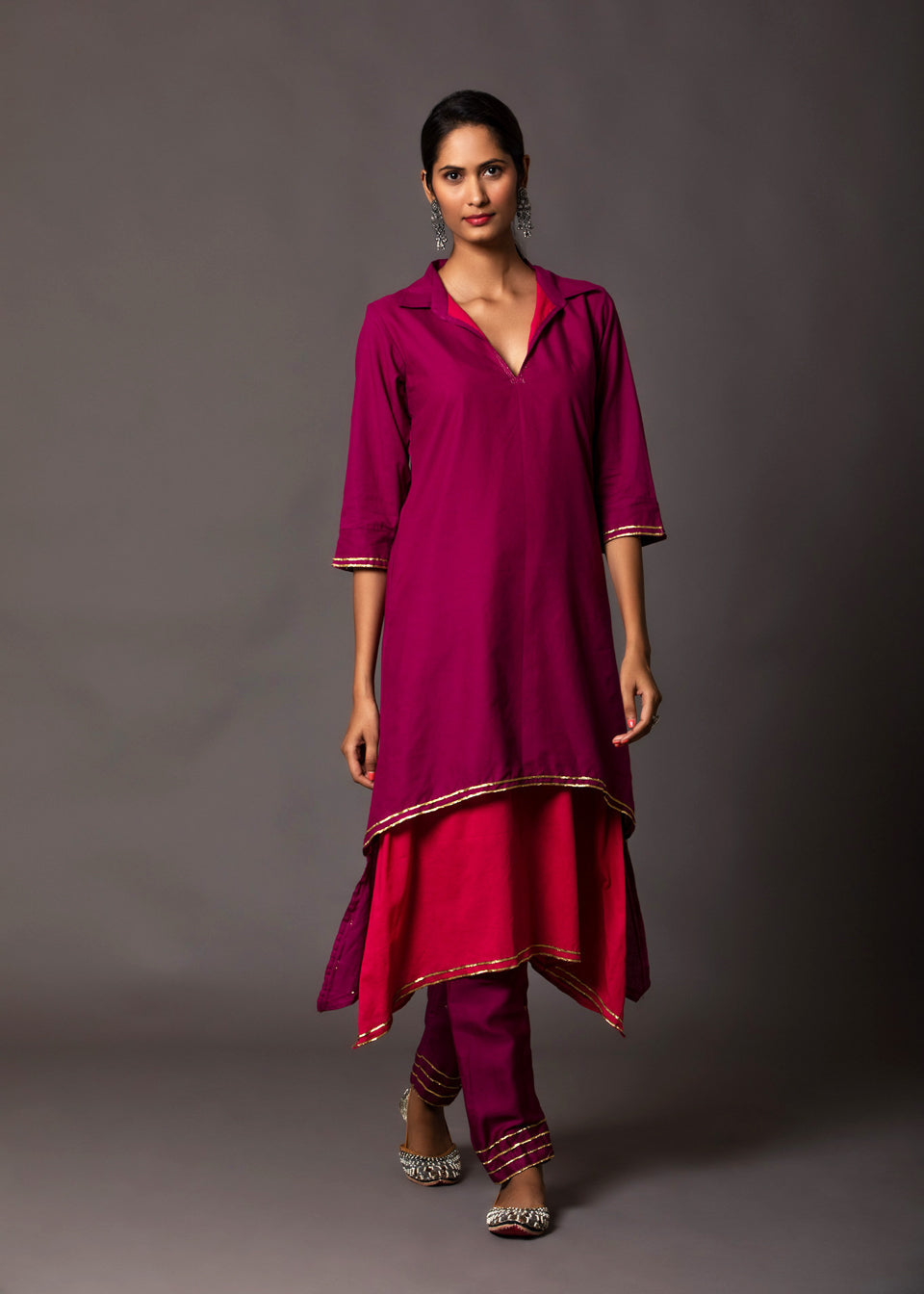 Woman wearing a layered magenta and pink shirt-style kurta with gold gota detailing and matching pants, styled with traditional juttis and silver earrings.

