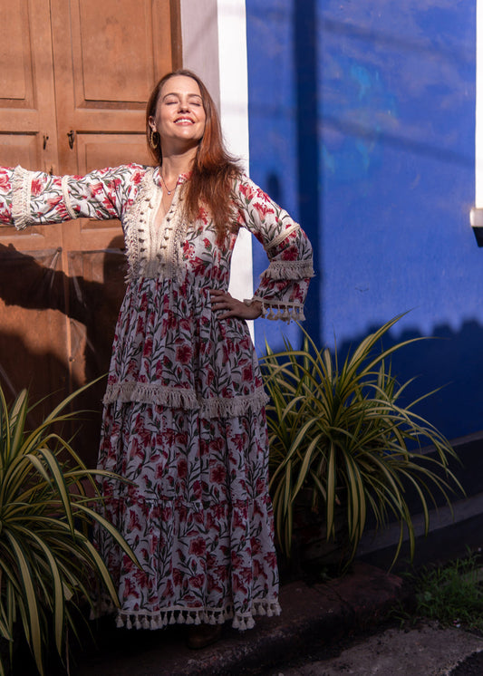 Woman wearing Mystical Garden boho floral maxi dress with lace and tassel detailing, standing against a blue wall in sunlight