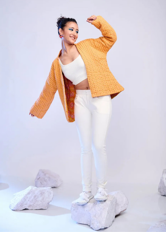 Model wearing an ochre yellow handquilted cotton jacket with multicolour threadwork and printed inner lining, styled over white leggings and sneakers on a minimalist set.