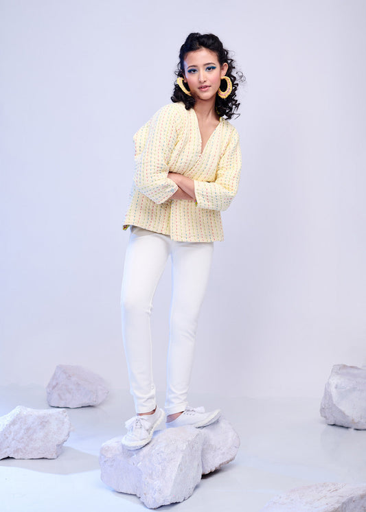 Model wearing a pastel yellow hand-quilted jacket with rainbow embroidery from The Silver Lining collection, styled with white pants and large statement earrings.
