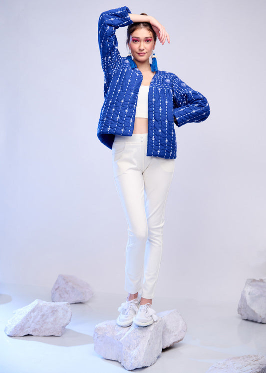 Model wearing a handquilted dark blue cotton jacket with white Katha embroidery, styled over white trousers and sneakers on a minimalist stone backdrop.