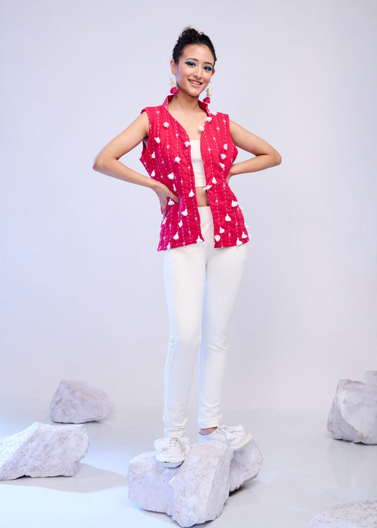 Model wearing a sleeveless red hand-quilted jacket with white floral embroidery over a white top and pants, standing playfully on textured rocks.


