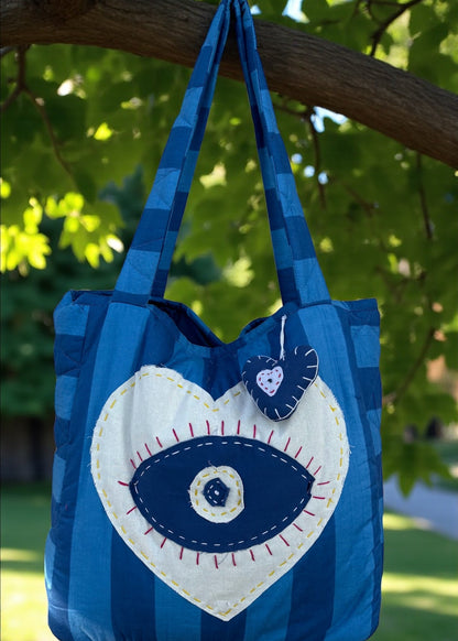 Blue striped quilted tote bag with heart-shaped evil-eye appliqué and matching padded charm hanging outdoors.